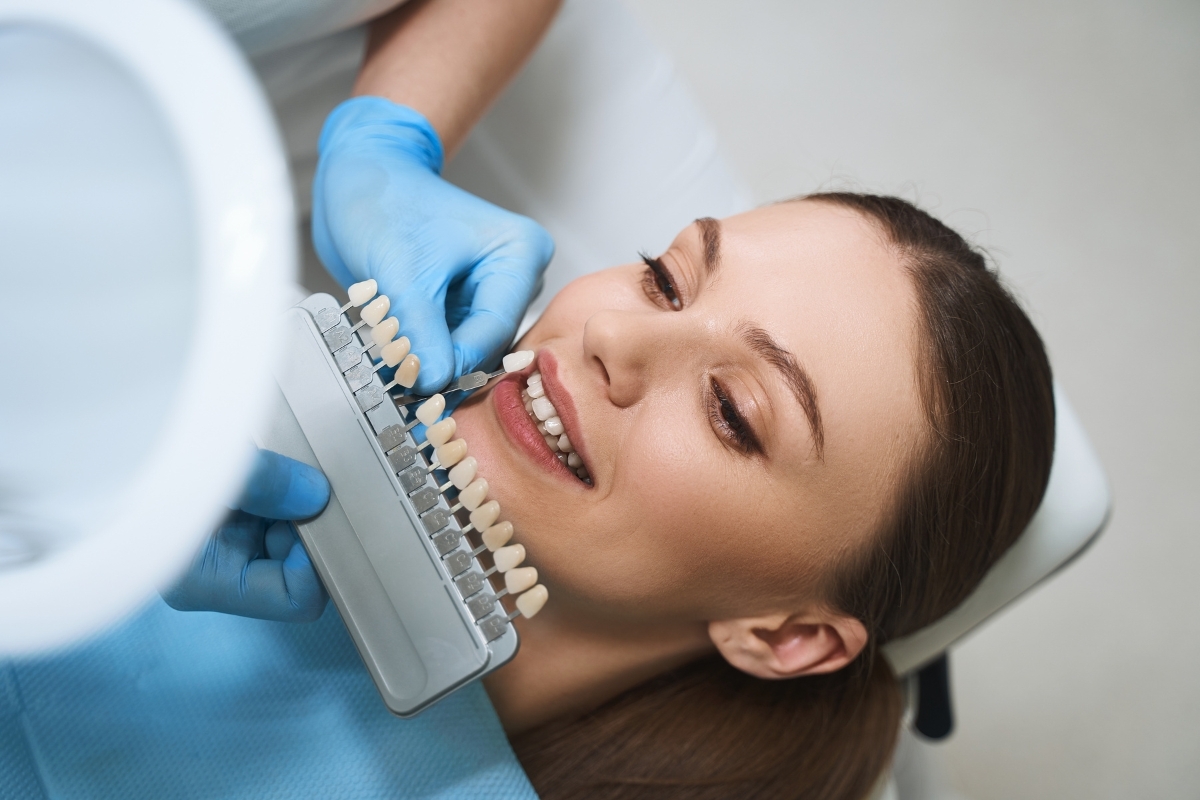 Dentist holding tooth shade guide next to patient's smile during veneer color matching consultation.